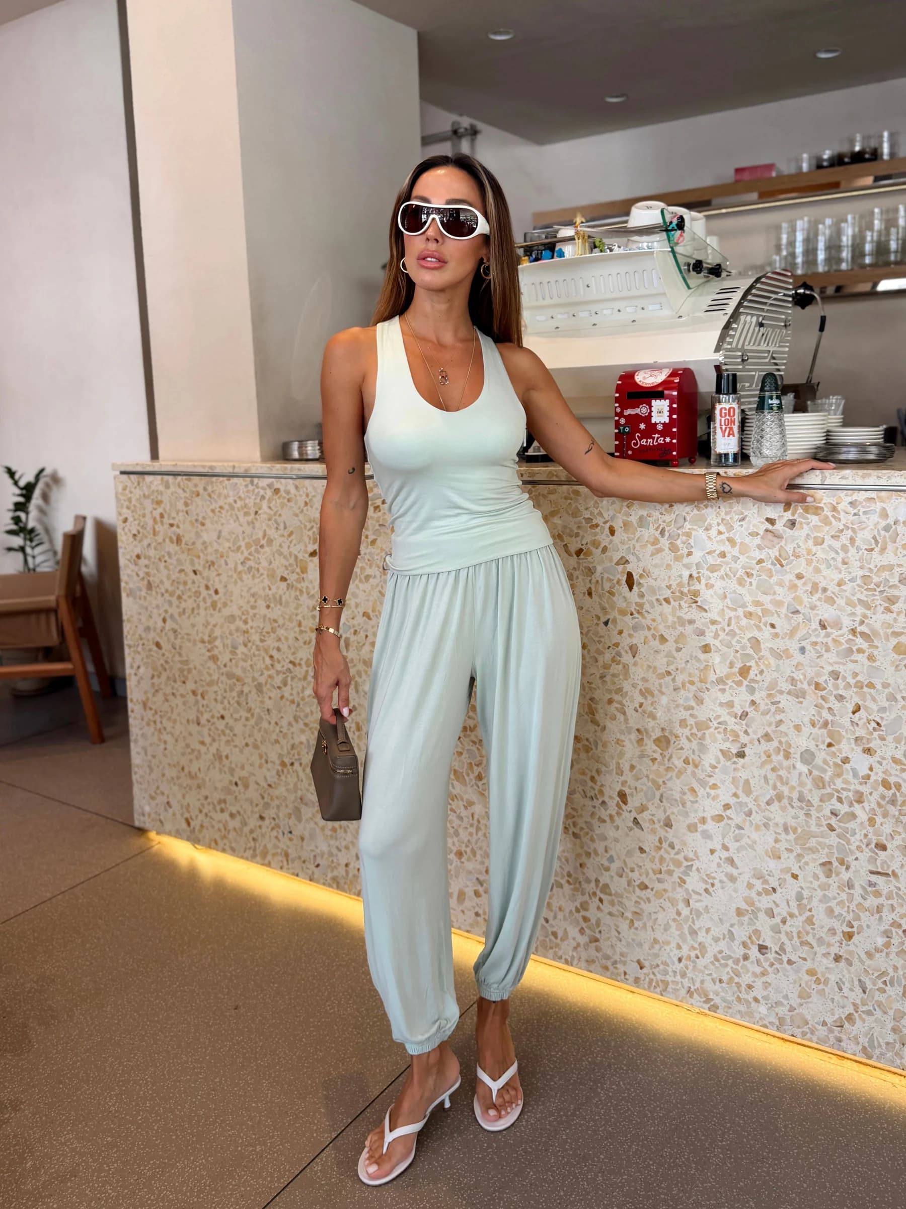 Auren Dual çift taraflı atlet matcha rengiyle: kafede terrazzo tezgâha yaslanan, güneş gözlüklü, matcha atlet ve aynı renk eşofman altı giyen, elinde çanta taşıyan model.