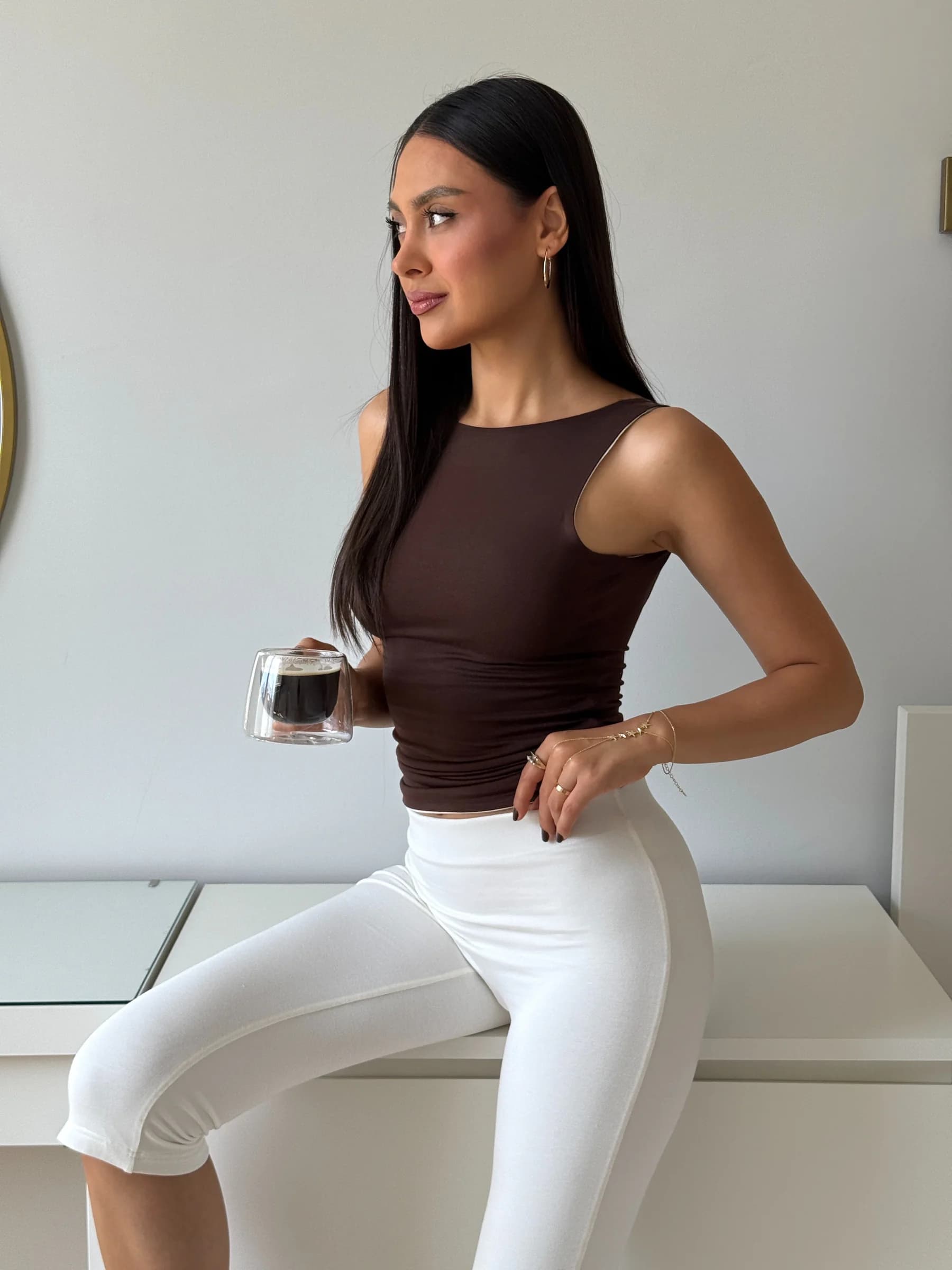 Deep Dual atletin chocolate yüzü önden: beyaz dolabın üzerinde oturan, elinde kahve bardağı tutan, beyaz kapri tayt giyen model.