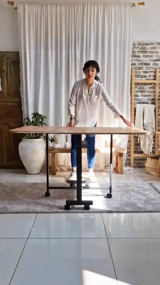 Massada katlanır masa videosundan bir kare: aydınlık bir odada beyaz gömlekli bir kadın tekerlekli ahşap katlanır masayı tamamen açmış, arkasında duruyor; arkada beyaz tül perde, tuğla duvar ve büyük vazoda bitki.