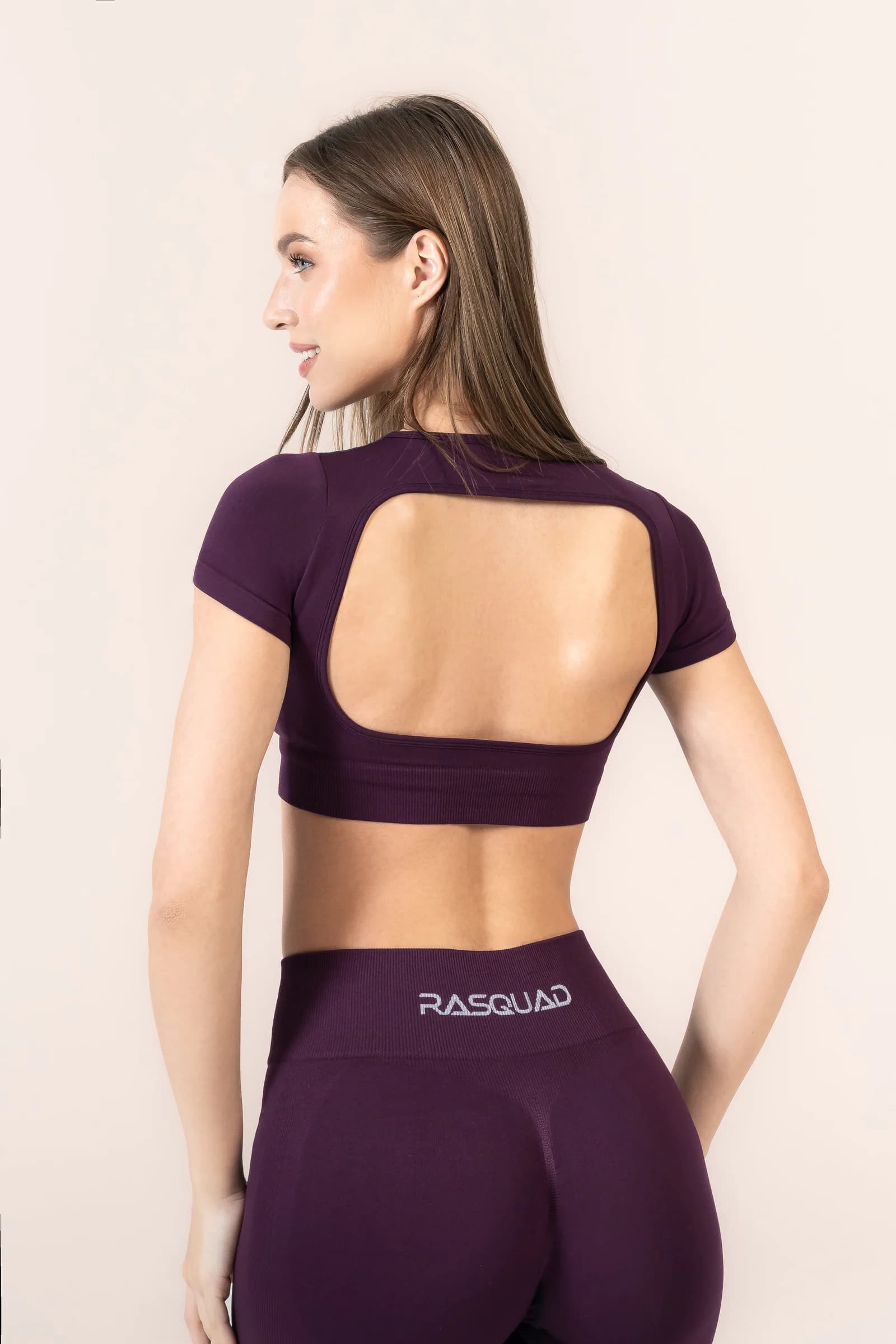 Sırtı açık crop top arkadan: açık gri zeminde mürdüm kısa kollu, sırtı oyuk crop top ve belinde RASQUAD yazılı mürdüm tayt giyen model.