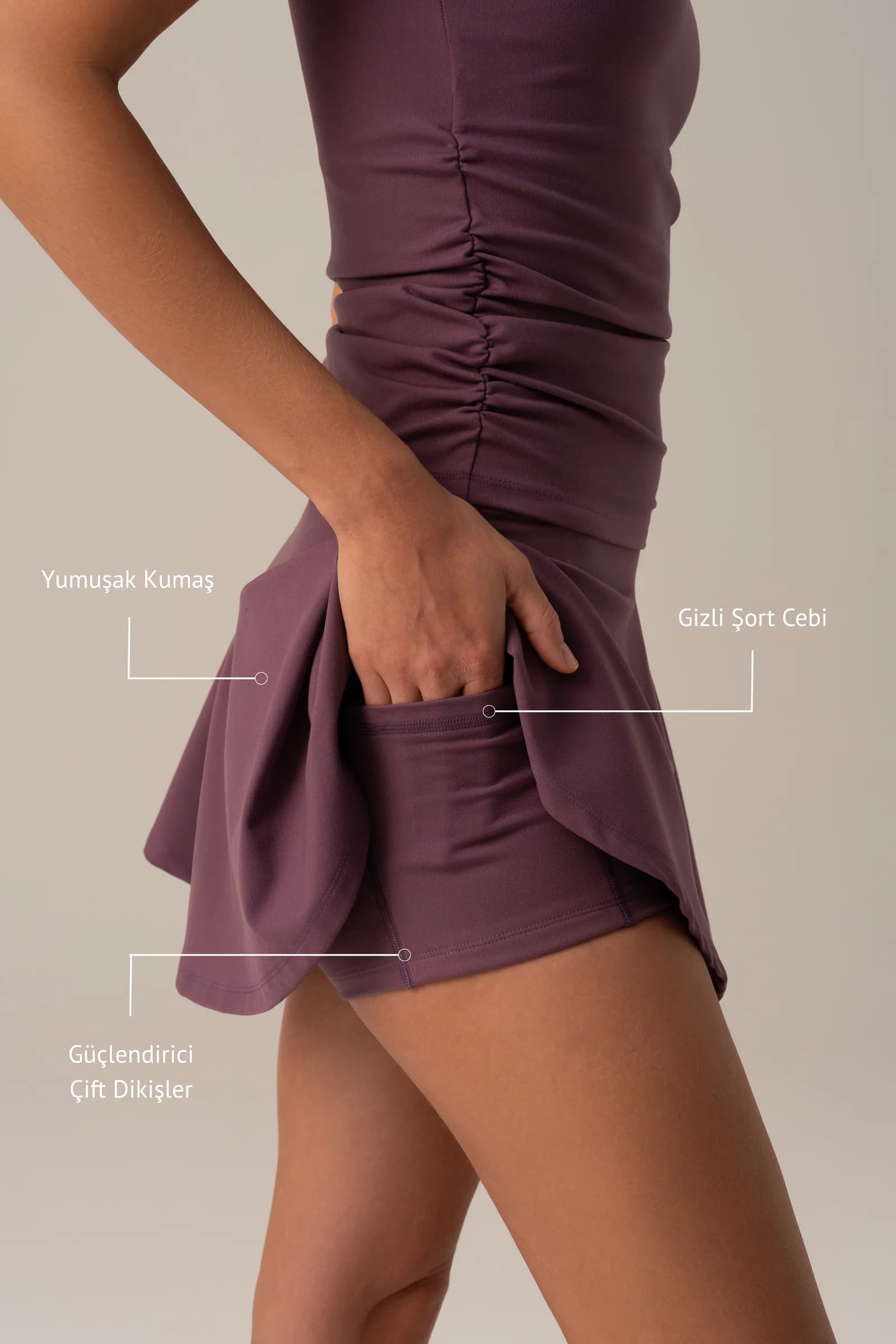 Match Point Şort Etek galerisinin notlu görseli: elini gizli cebe sokmuş, mürdüm şort etek giyen modelin yandan yakın planı; fotoğraf üzerinde Yumuşak Kumaş, Gizli Şort Cebi ve Güçlendirici Çift Dikişler notları.